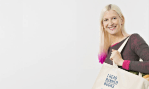 A smiling woman with blonde hair featuring pink ombre ends holds a canvas tote bag over her shoulder. She wears a dark long-sleeved athletic top with pink accents. The cream-colored tote bag displays the text "I READ BANNED BOOKS" along with what appears to be a library card design listing various book titles. The image is set against a plain light background, promoting literary freedom and the importance of accessing diverse literature. The overall aesthetic conveys a positive, advocacy-focused message about reading and intellectual freedom.