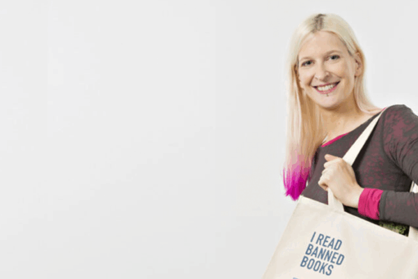 A smiling woman with blonde hair featuring pink ombre ends holds a canvas tote bag over her shoulder. She wears a dark long-sleeved athletic top with pink accents. The cream-colored tote bag displays the text "I READ BANNED BOOKS" along with what appears to be a library card design listing various book titles. The image is set against a plain light background, promoting literary freedom and the importance of accessing diverse literature. The overall aesthetic conveys a positive, advocacy-focused message about reading and intellectual freedom.