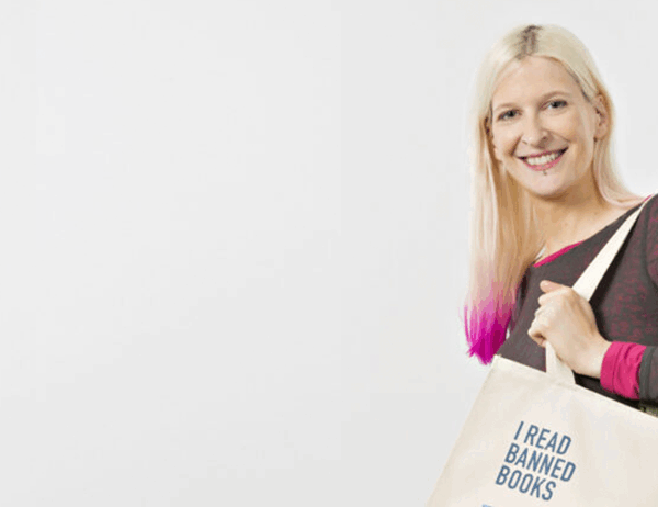A smiling woman with blonde hair featuring pink ombre ends holds a canvas tote bag over her shoulder. She wears a dark long-sleeved athletic top with pink accents. The cream-colored tote bag displays the text "I READ BANNED BOOKS" along with what appears to be a library card design listing various book titles. The image is set against a plain light background, promoting literary freedom and the importance of accessing diverse literature. The overall aesthetic conveys a positive, advocacy-focused message about reading and intellectual freedom.
