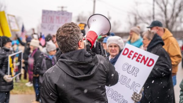 A person in a black jacket speaks through a megaphone at an outdoor protest rally during light snowfall. Behind them, demonstrators hold signs including one that reads "DEFEND OUR PUBLIC WORKERS" in red lettering. The crowd includes people of various ages wearing winter clothing like hats and coats. Bare trees and buildings are visible in the background. The scene captures civic engagement and collective action on a cold, overcast day, with protesters gathered to advocate for labor rights and workers' protections.