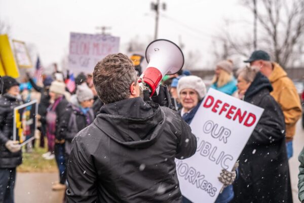 A person in a black jacket speaks through a megaphone at an outdoor protest rally during light snowfall. Behind them, demonstrators hold signs including one that reads "DEFEND OUR PUBLIC WORKERS" in red lettering. The crowd includes people of various ages wearing winter clothing like hats and coats. Bare trees and buildings are visible in the background. The scene captures civic engagement and collective action on a cold, overcast day, with protesters gathered to advocate for labor rights and workers' protections.