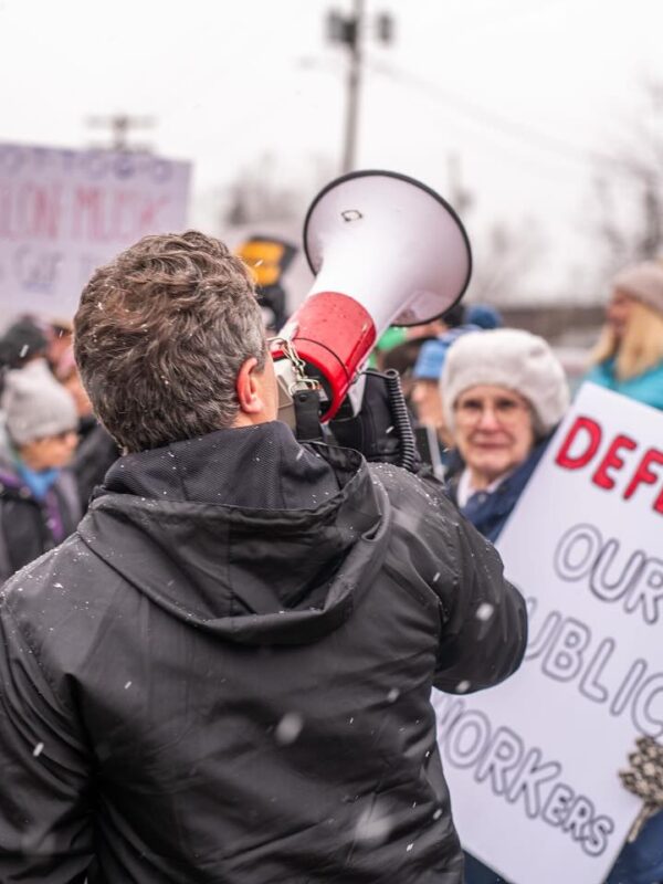 A person in a black jacket speaks through a megaphone at an outdoor protest rally during light snowfall. Behind them, demonstrators hold signs including one that reads "DEFEND OUR PUBLIC WORKERS" in red lettering. The crowd includes people of various ages wearing winter clothing like hats and coats. Bare trees and buildings are visible in the background. The scene captures civic engagement and collective action on a cold, overcast day, with protesters gathered to advocate for labor rights and workers' protections.