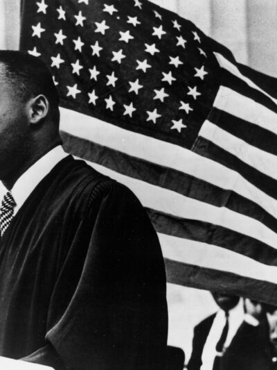 Black and White Photo shows Dr. Martin Luther King Jr. in front of an American flag