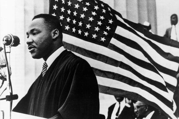 Black and White Photo shows Dr. Martin Luther King Jr. in front of an American flag