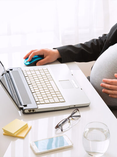 A pregnant worker touches their belly while performing work duties on a laptop