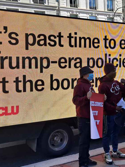 Protestors stand in front of a digital sign that says "It's past time to end Trump-era policies at the border"