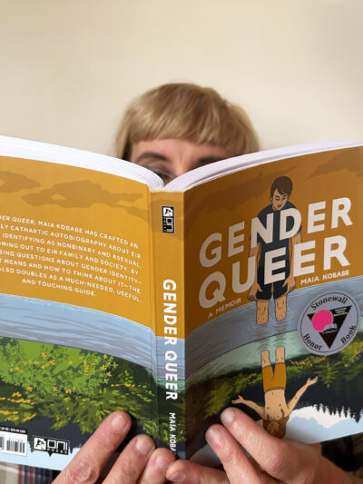 A white person with blond hair and glasses reads a copy of "Gender Queer" against a solid white backdrop