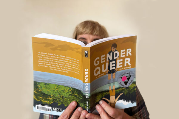 A white person with blond hair and glasses reads a copy of "Gender Queer" against a solid white backdrop