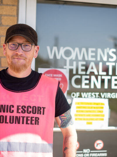 Rusty Williams wears a pink vest reading Clinic Escort Volunteer in front of the entrance to the Women's Health Center of West Virginia