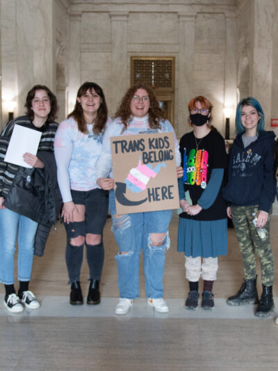 Seven attendees of the Public Hearing on HB 2007 pose with a sign that reads "Trans Kids Belong Here and a picture of West Virginia"