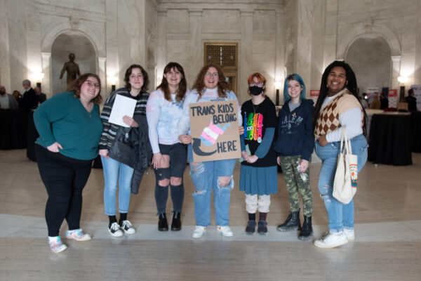 Seven attendees of the Public Hearing on HB 2007 pose with a sign that reads "Trans Kids Belong Here and a picture of West Virginia"