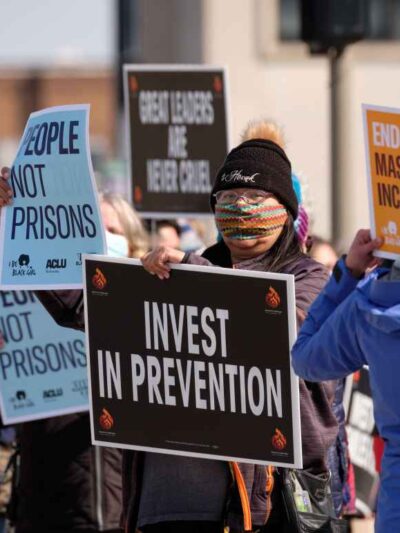 Protesters wave signs saying "INVEST IN PREVENTION," "PEOPLE NOT PRISONS," AND "END MASS INCARCERATION."