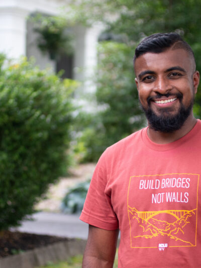 Kasun Wijayagurusinghe is pictured in an orange Build Bridges Not Walls tee shirt in front of the ACLU-WV Charleston Office