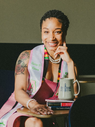 Tiara Rowe is a young Black women with short hair and a shoulder tattoo sits at a table with books and a coffee mug