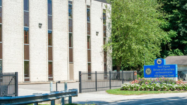 A blue sign with gold text displays the WV Division of Correction of Rehabilitation outside of the parking lot for the building. The building is off-white with long, vertical windows and is surrounded by a black metal fence.
