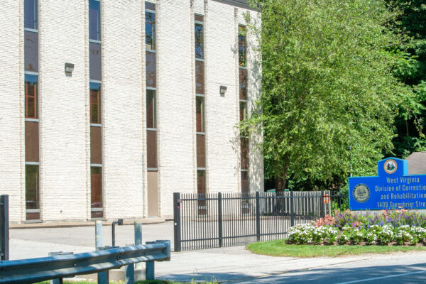 A blue sign with gold text displays the WV Division of Correction of Rehabilitation outside of the parking lot for the building. The building is off-white with long, vertical windows and is surrounded by a black metal fence.