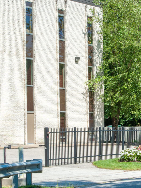 A blue sign with gold text displays the WV Division of Correction of Rehabilitation outside of the parking lot for the building. The building is off-white with long, vertical windows and is surrounded by a black metal fence.