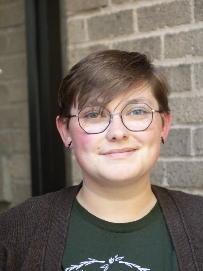 Sam Green is a white person with short medium brown hair, blue eyes and glasses, wearing a green shirt and brown sweater, standing outside of the ACLU offices.