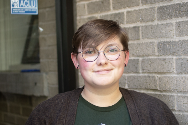 Sam Green is a white person with short medium brown hair, blue eyes and glasses, wearing a green shirt and brown sweater, standing outside of the ACLU offices.