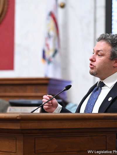 Eli Baumwell speaks at the West Virginia Legislature wearing a dark suit and blue tie.
