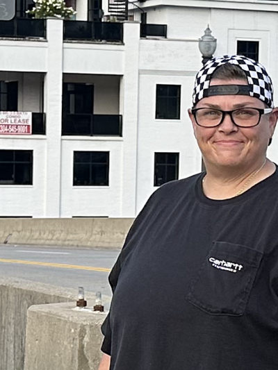 Miranda Johnson, a white woman with dark glasses, short hair and a backwards checker-print baseball hat, smiles for the camera with downtown Charleston's waterfront in the background.