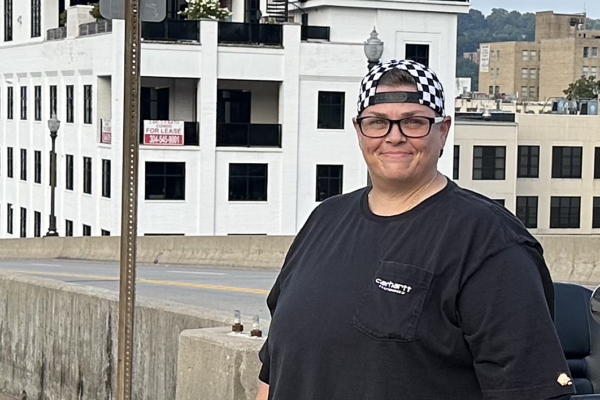 Miranda Johnson, a white woman with dark glasses, short hair and a backwards checker-print baseball hat, smiles for the camera with downtown Charleston's waterfront in the background.