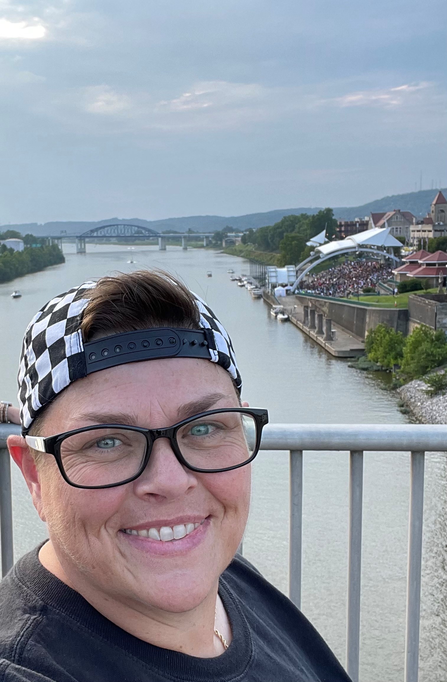 Miranda Johnson, a white woman with dark glasses, short hair and a backwards checker-print baseball hat, smiles for the camera with downtown Charleston's waterfront in the background.