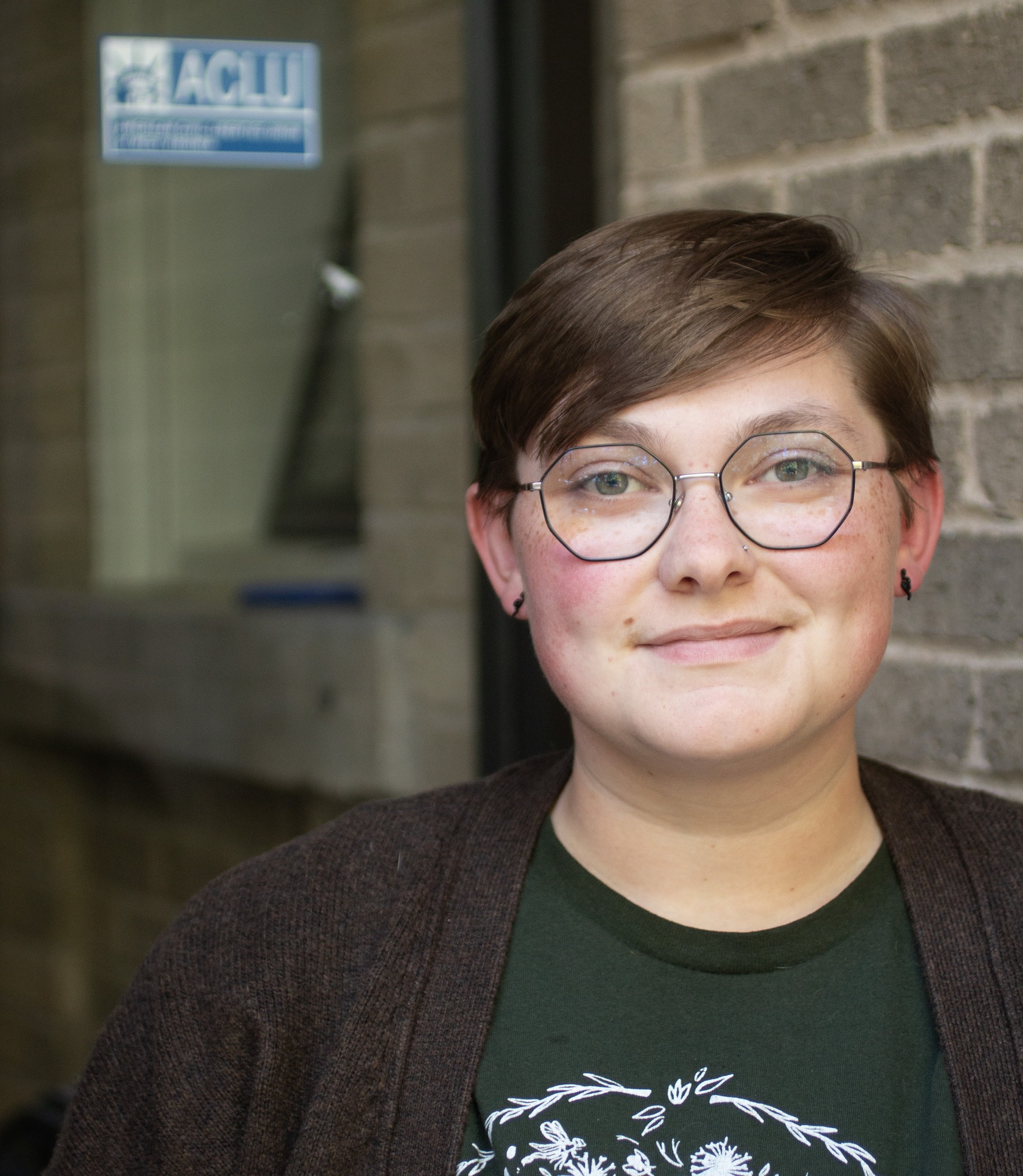 Sam Green, a white person with light brown hair and glasses is pictured in front of a glass door with a blue ACLU sticker on it