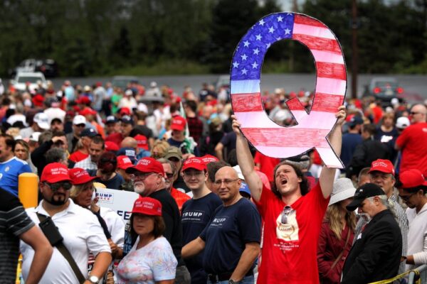 A white man with long hair hold up a large "Q" painted like the American flag for the conspiracy theory Q Anon.