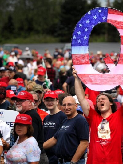 A white man with long hair hold up a large "Q" painted like the American flag for the conspiracy theory Q Anon.