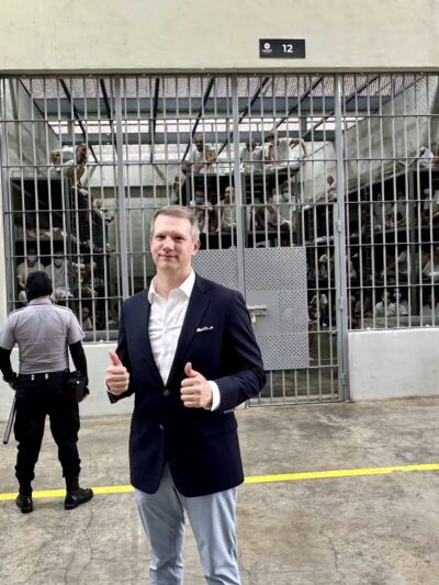 Riley Moore, a Congressman, stands in a suit giving a thumbs up in front of a large prison cell holding numerous people being watched over by a guard