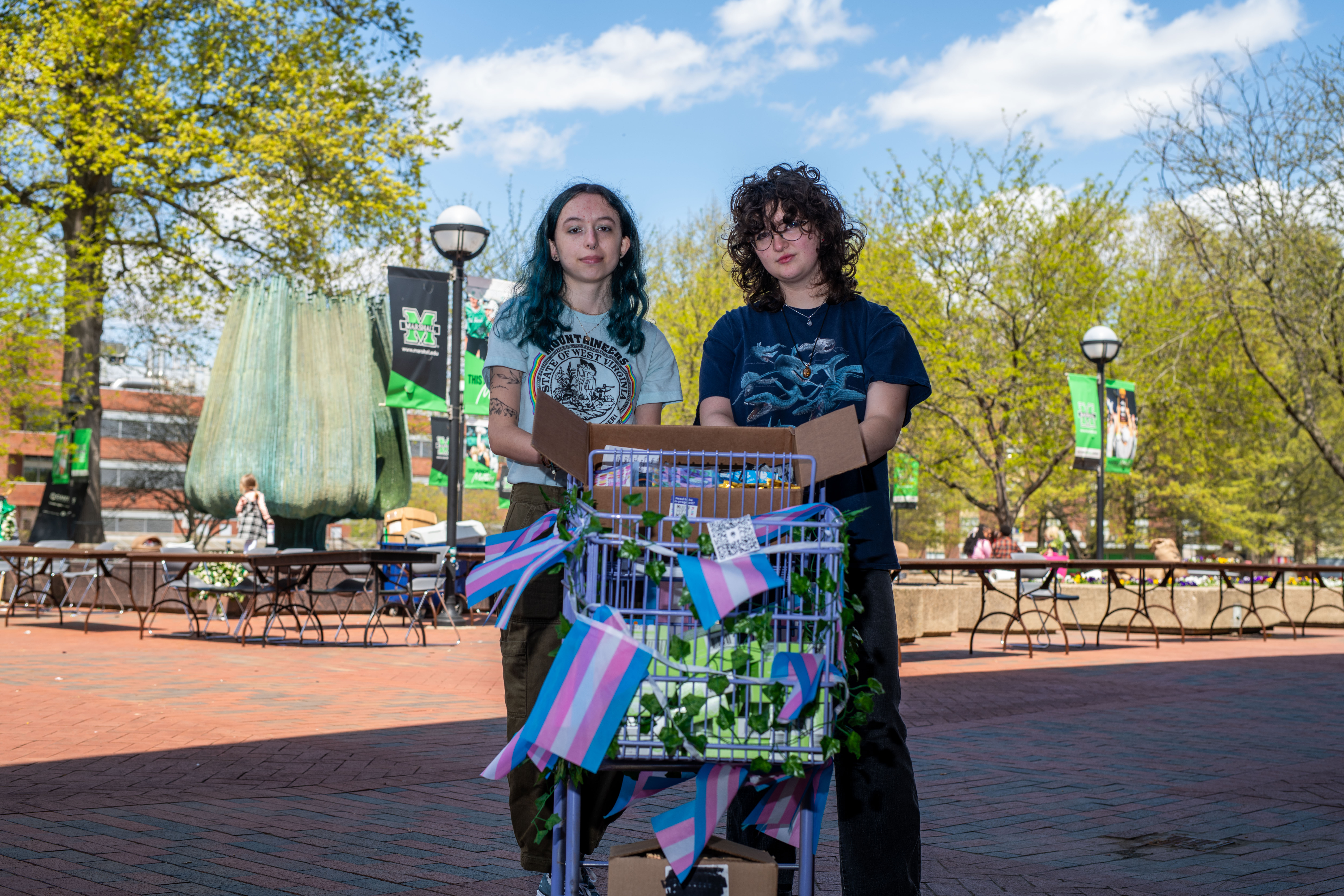 Two people stand behind a shopping cart decorated with pink, blue, and white transgender pride flags and green vines. The cart is filled with boxes, and the scene is outdoors on a brick plaza with trees, banners, and a large fountain nearby.