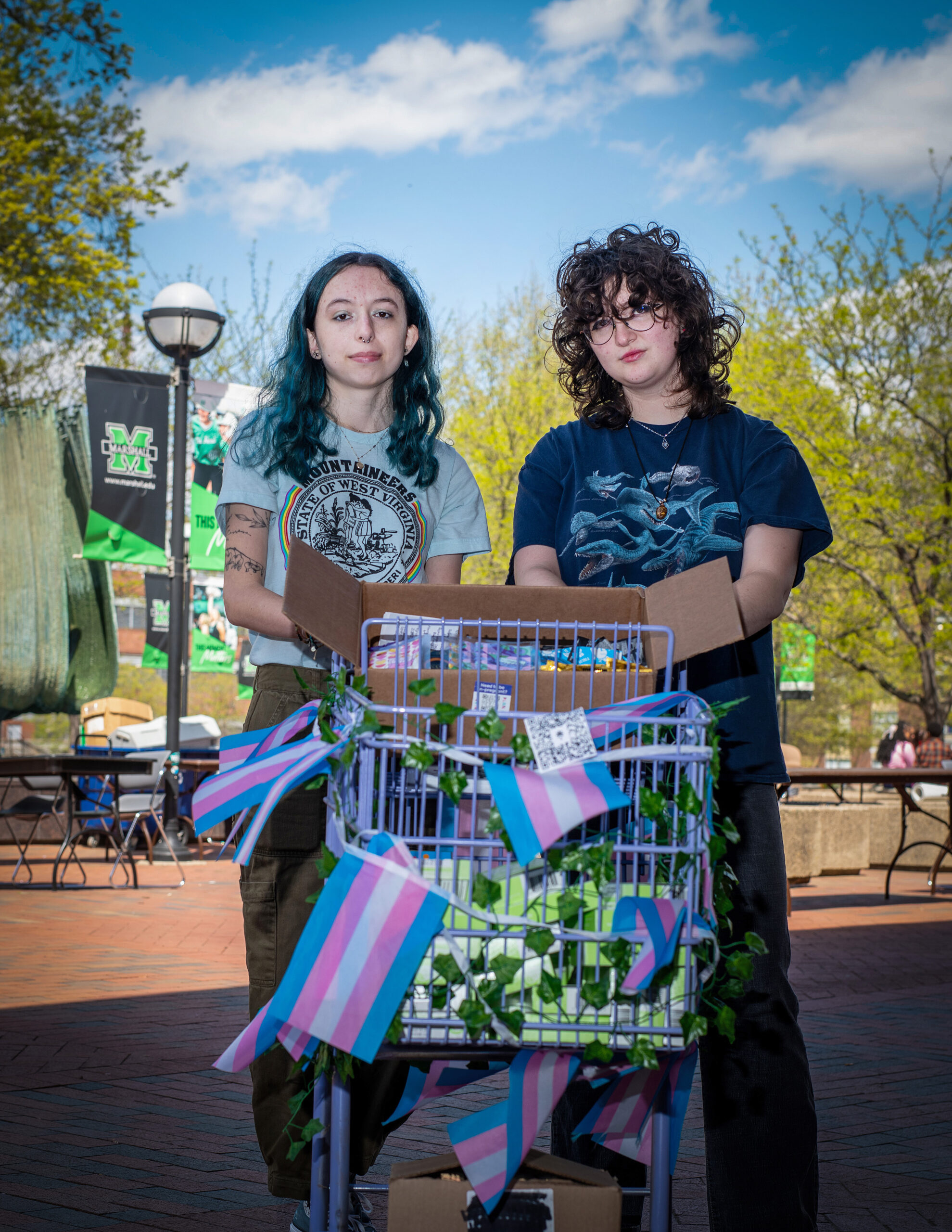 Two young people standing behind shopping cart decorated with blue, pink and white striped ribbons and green foliage on college campus plaza, with university signage visible in background under sunny sky.