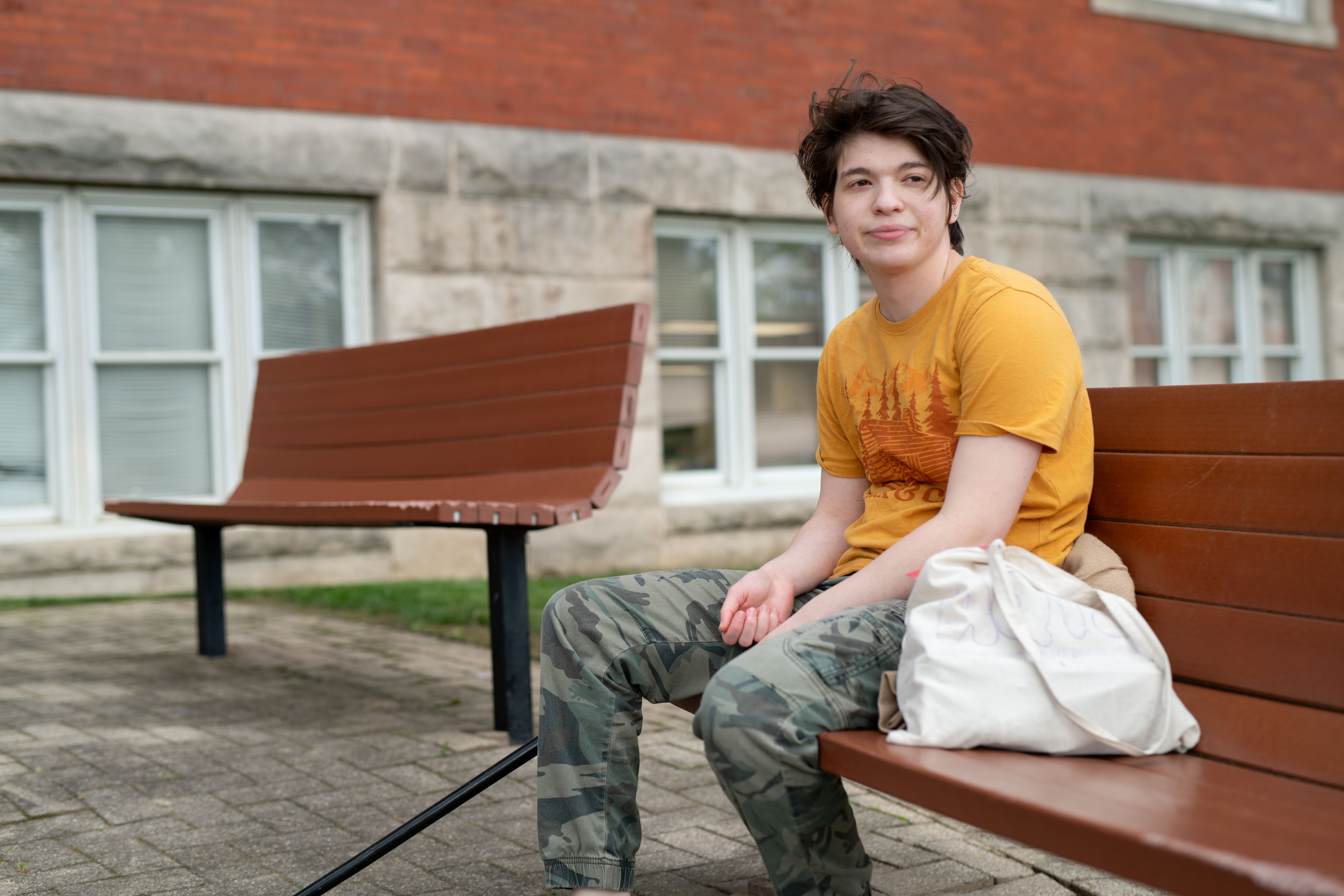 Young person with short brown hair in yellow graphic t-shirt and camouflage pants sitting on wooden bench with white bag, in front of brick and white building with multiple windows.
