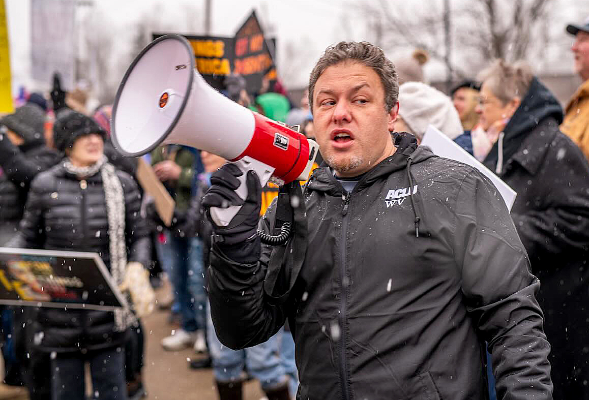 Person in dark jacket speaking into red and white megaphone at outdoor winter gathering with crowd and falling snow visible in background.