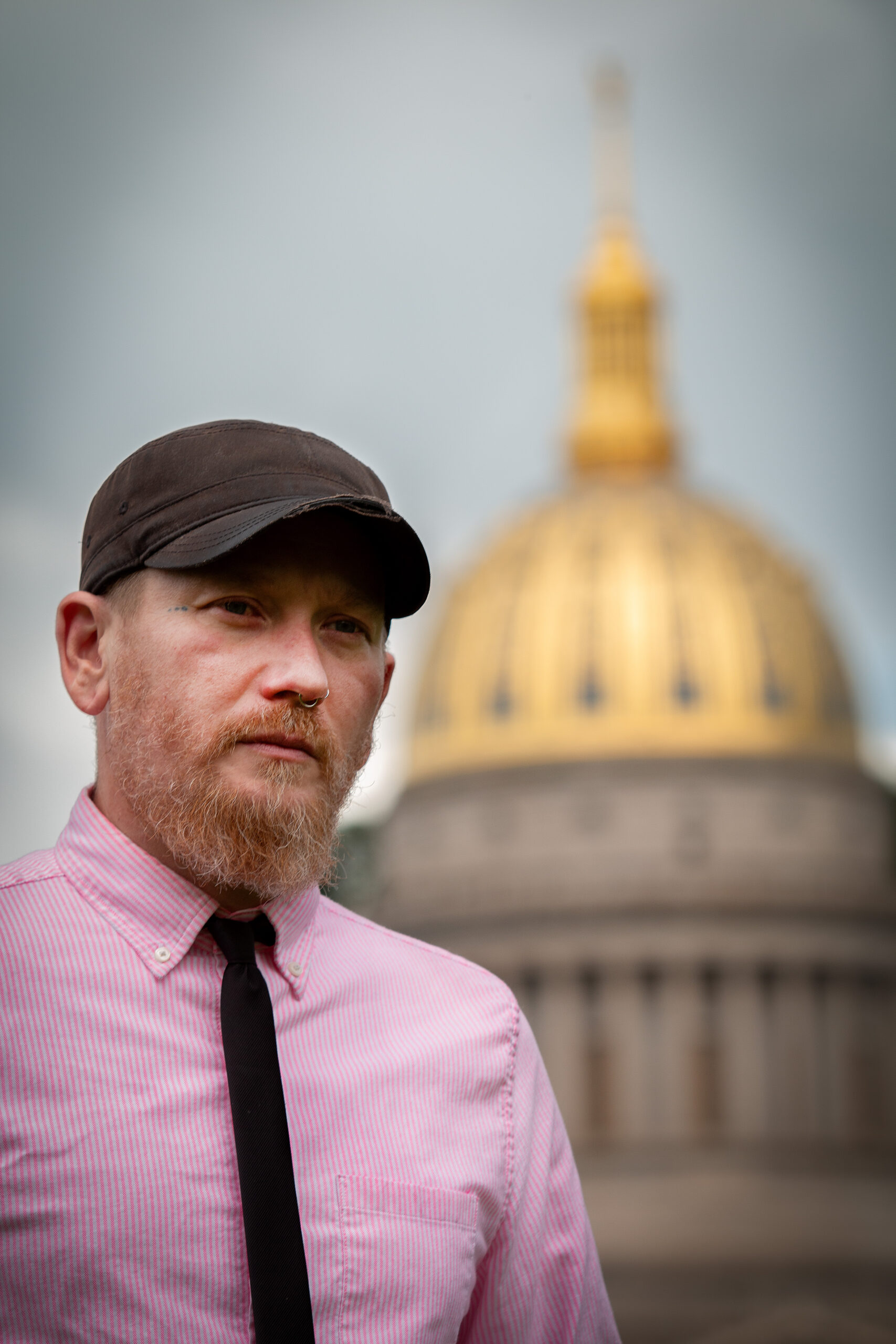 Bearded man in brown cap, pink striped dress shirt and black tie standing in foreground with blurred golden-domed government building or capitol dome visible in background on overcast day.