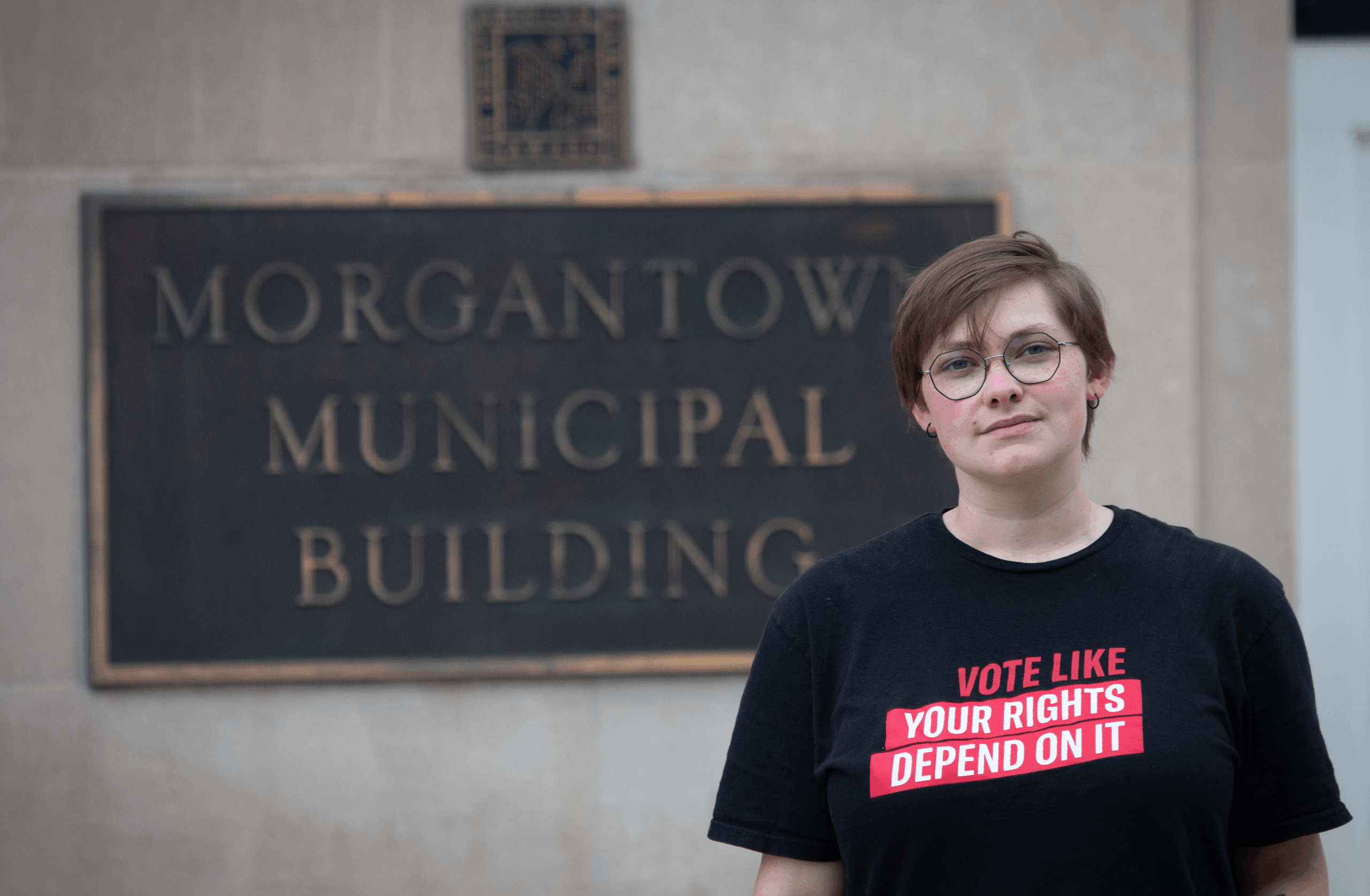 Person wearing glasses and black t-shirt with 'Vote Like Your Rights Depend On It' message standing in front of Morgantown Municipal Building sign.