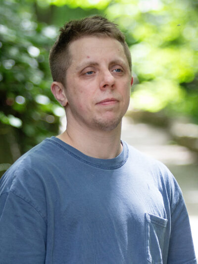 Robb Livingood, a white man with short light brown hair and a tattoo sleeve on his left arm, is pictured in a light blue shirt standing along a nature trail.