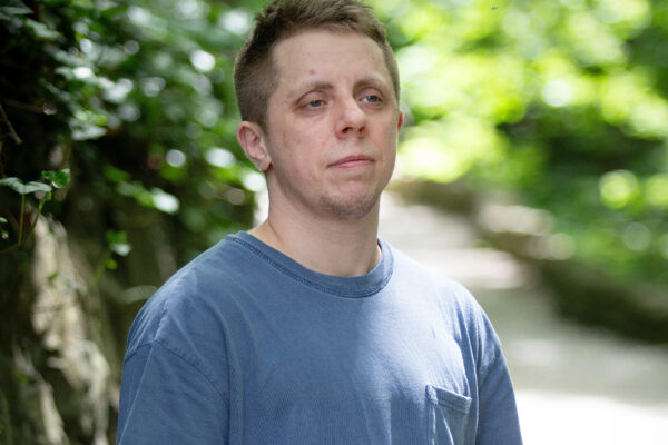 Robb Livingood, a white man with short light brown hair and a tattoo sleeve on his left arm, is pictured in a light blue shirt standing along a nature trail.