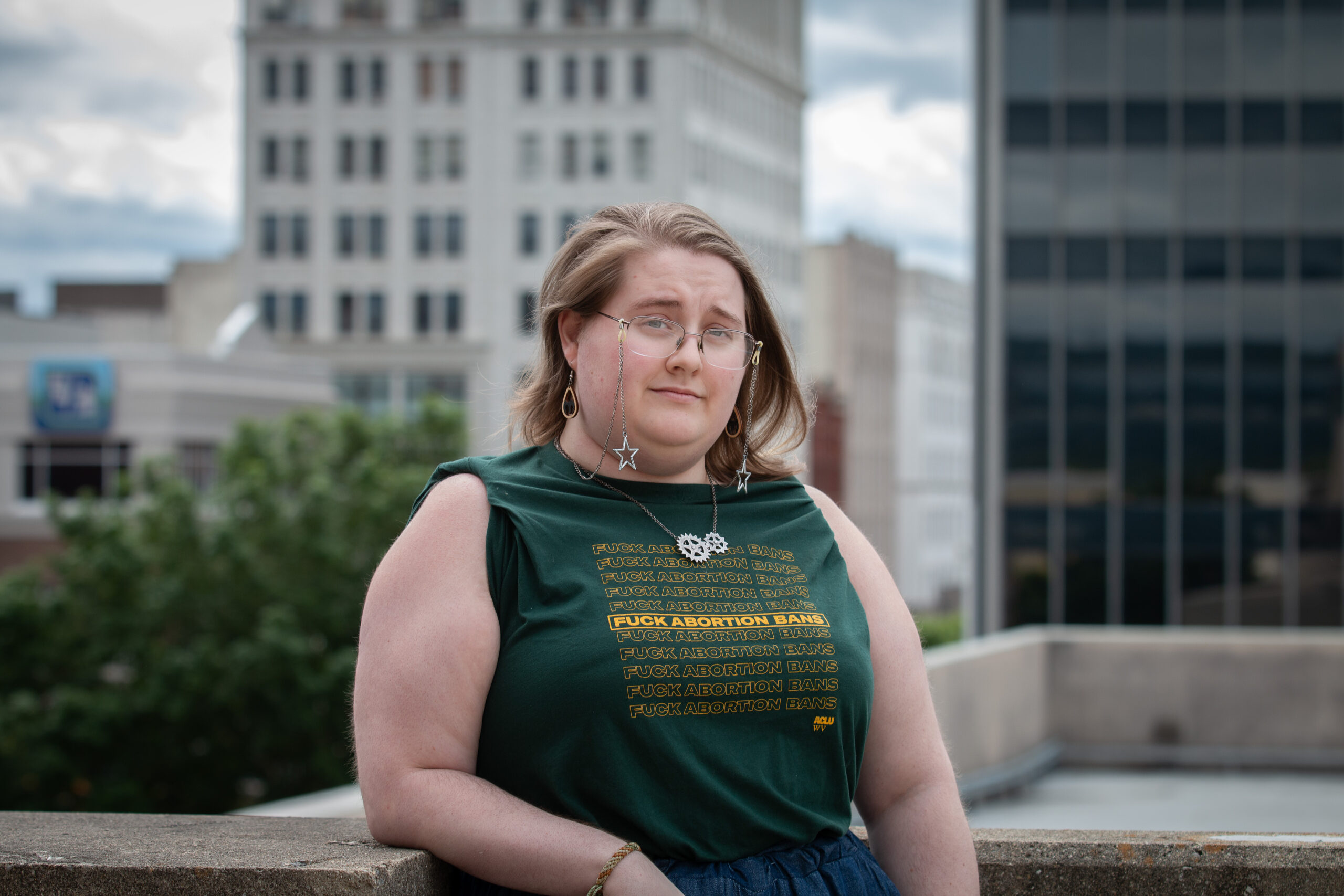 Young person with glasses and blonde hair in green tank top and blue wide-leg pants holding orange walking cane, standing on urban rooftop or plaza with tall office buildings and parking meters visible in background.