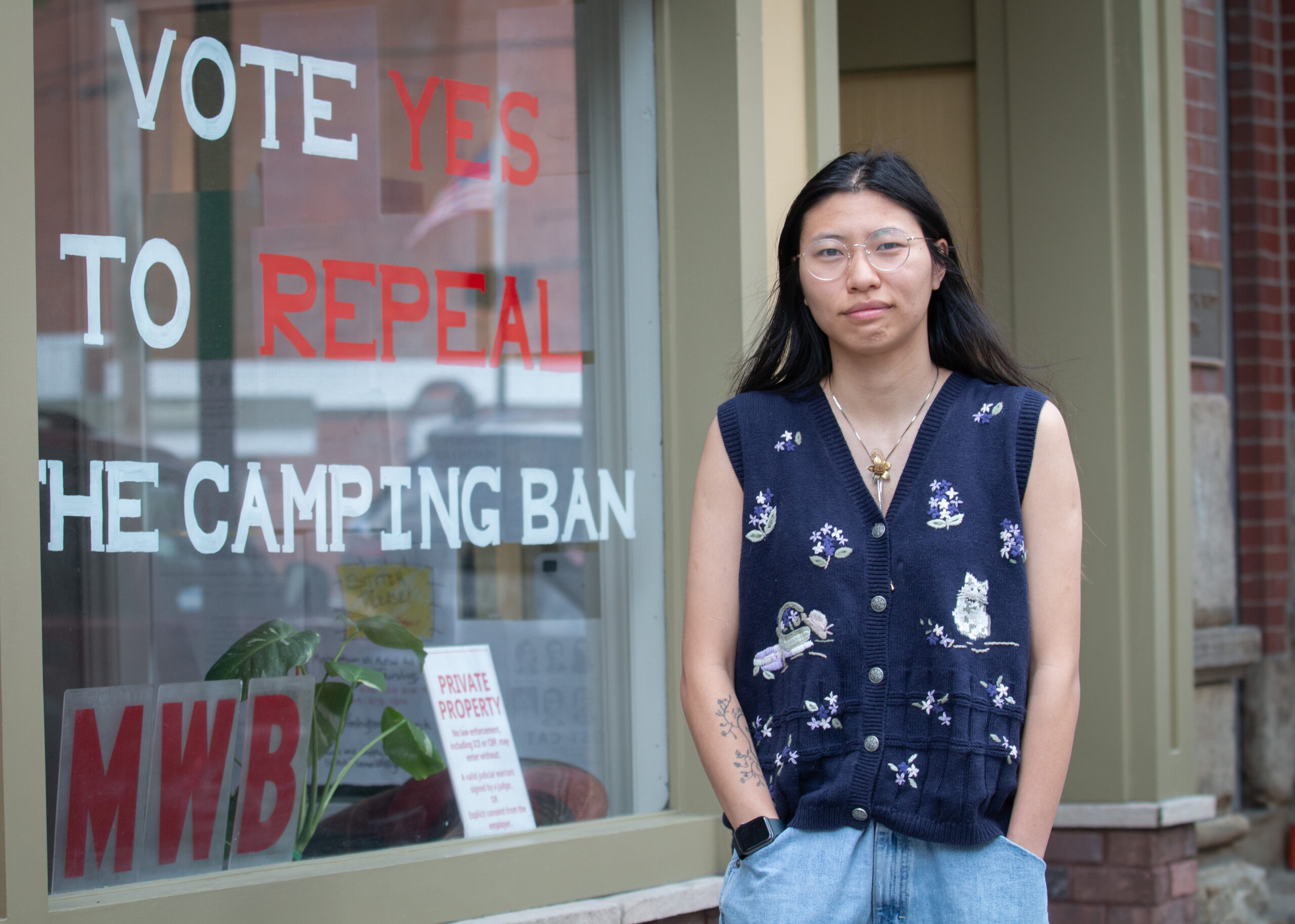 A young person in a navy floral cardigan and glasses standing in front of storefront window displaying