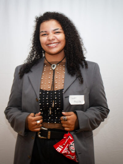 A smiling person with long curly black hair wearing a gray blazer over a brown and black polka-dot blouse stands against a light gray backdrop. They wear a name tag and hold what appears to be a small notebook or plaque, with a red bandana visible at their waist.