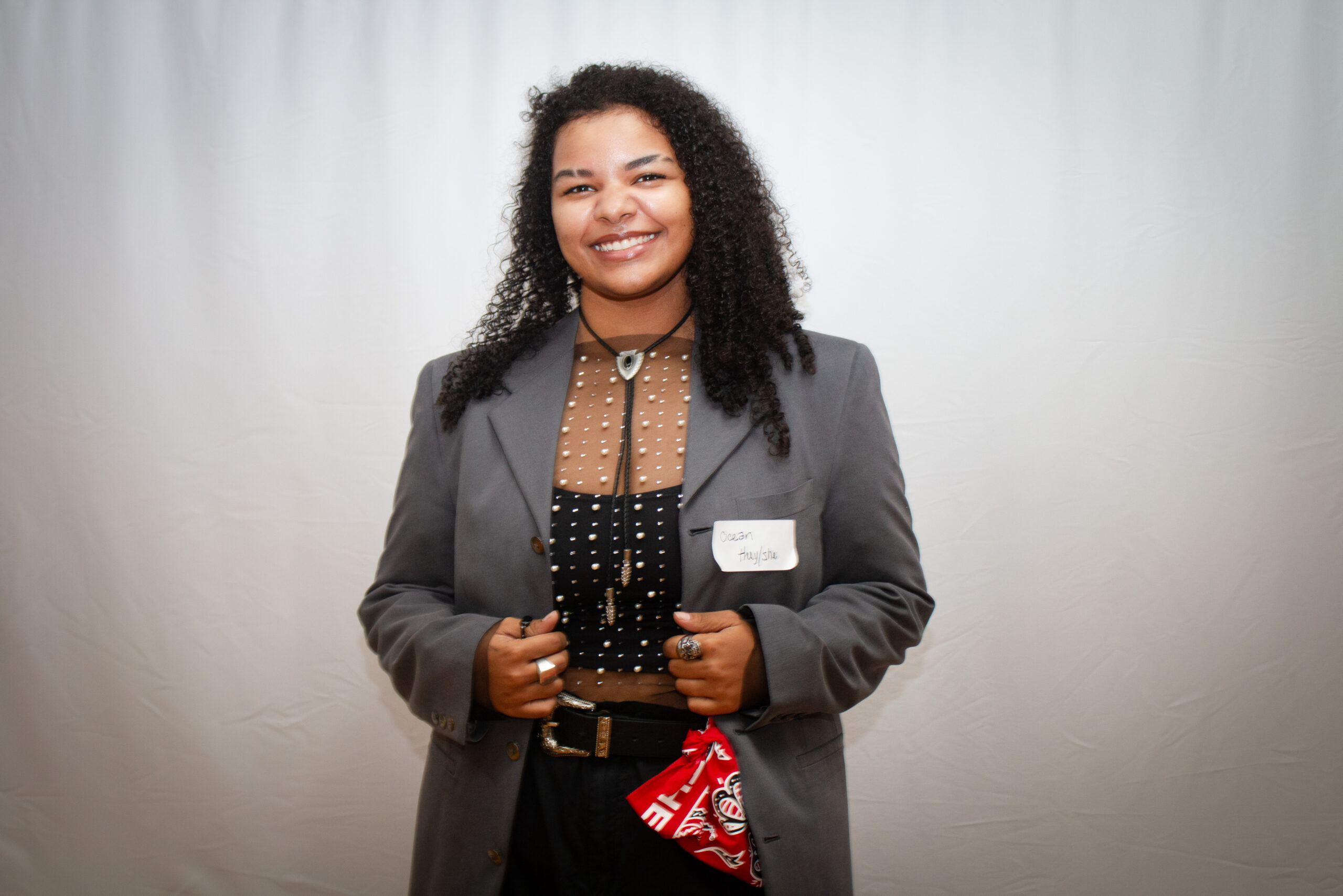 A smiling person with long curly black hair wearing a gray blazer over a brown and black polka-dot blouse stands against a light gray backdrop. They wear a name tag and hold what appears to be a small notebook or plaque, with a red bandana visible at their waist.