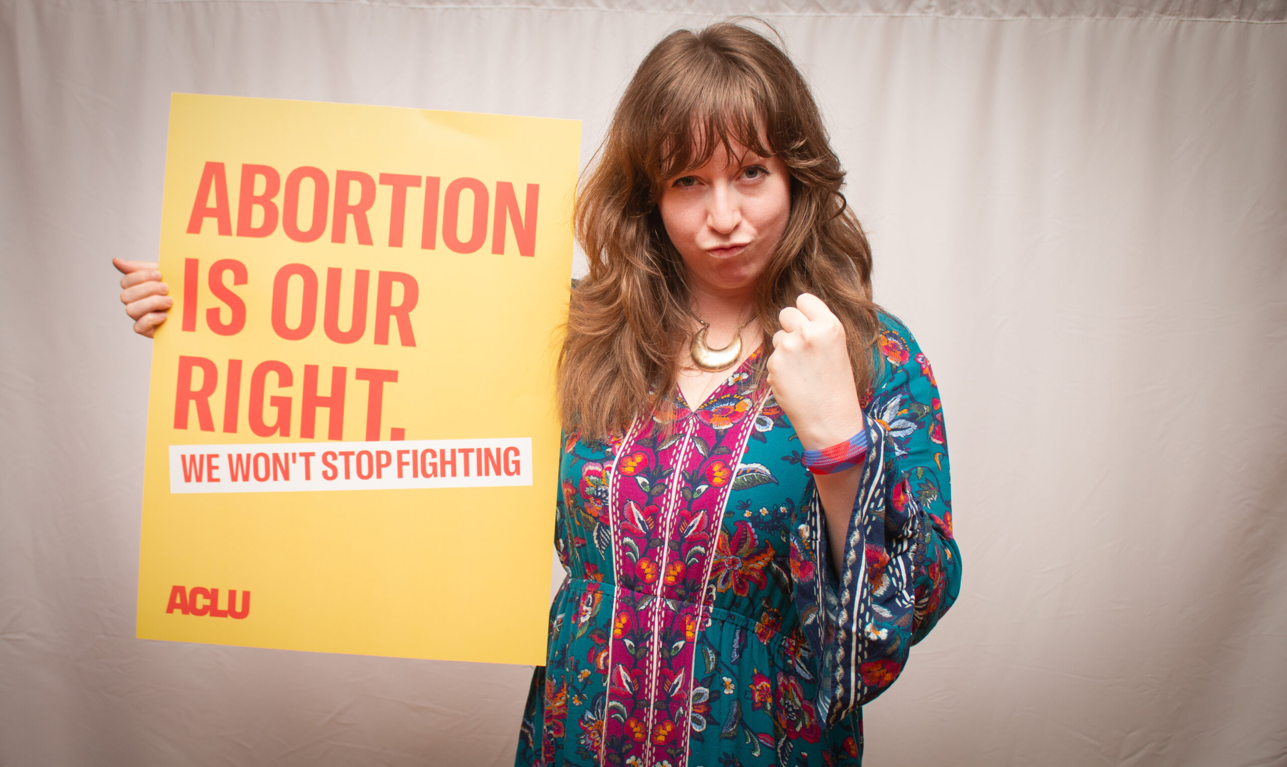 A person wearing a vibrant floral-patterned dress in turquoise, pink, and multicolored tones holds a yellow ACLU sign reading "ABORTION IS OUR RIGHT. WE WON'T STOP FIGHTING." They stand with one fist raised in a determined pose against a light backdrop, wearing a crescent moon necklace.