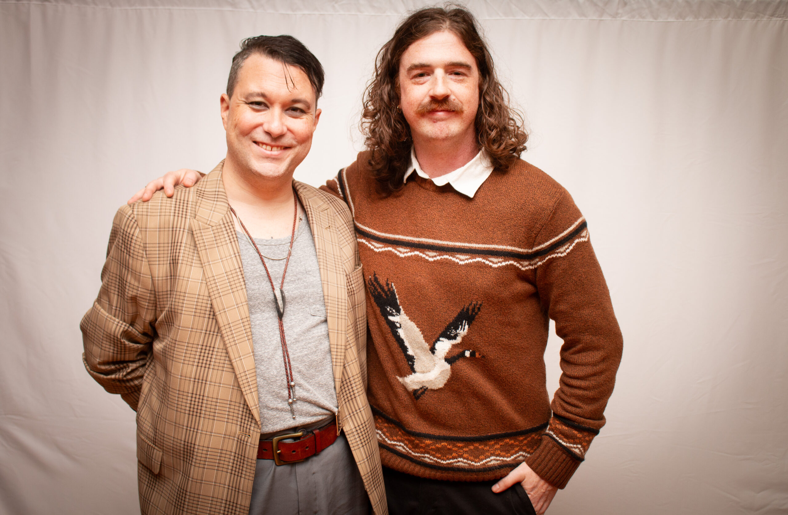 Two people stand close together smiling at the camera against a light backdrop. The person on left wears a tan plaid blazer with gray shirt, while the person on right wears a rust-colored sweater with a white collar and decorative bird pattern, sporting a mustache and shoulder-length wavy hair.
