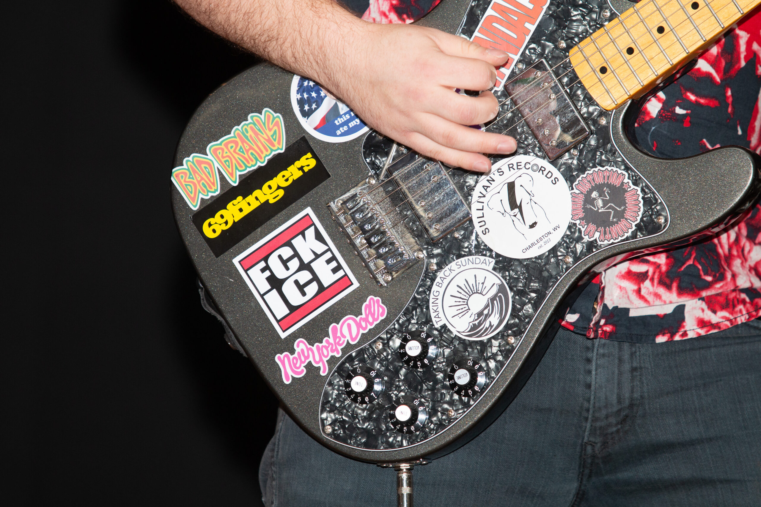 Close-up of hands playing an electric guitar covered in various stickers and decals, including "Bad Brains," "Godfinger," "FCK ICE," "Sullivan's Records," and other band and venue stickers. The guitar has a sparkly black pickguard and maple fretboard.