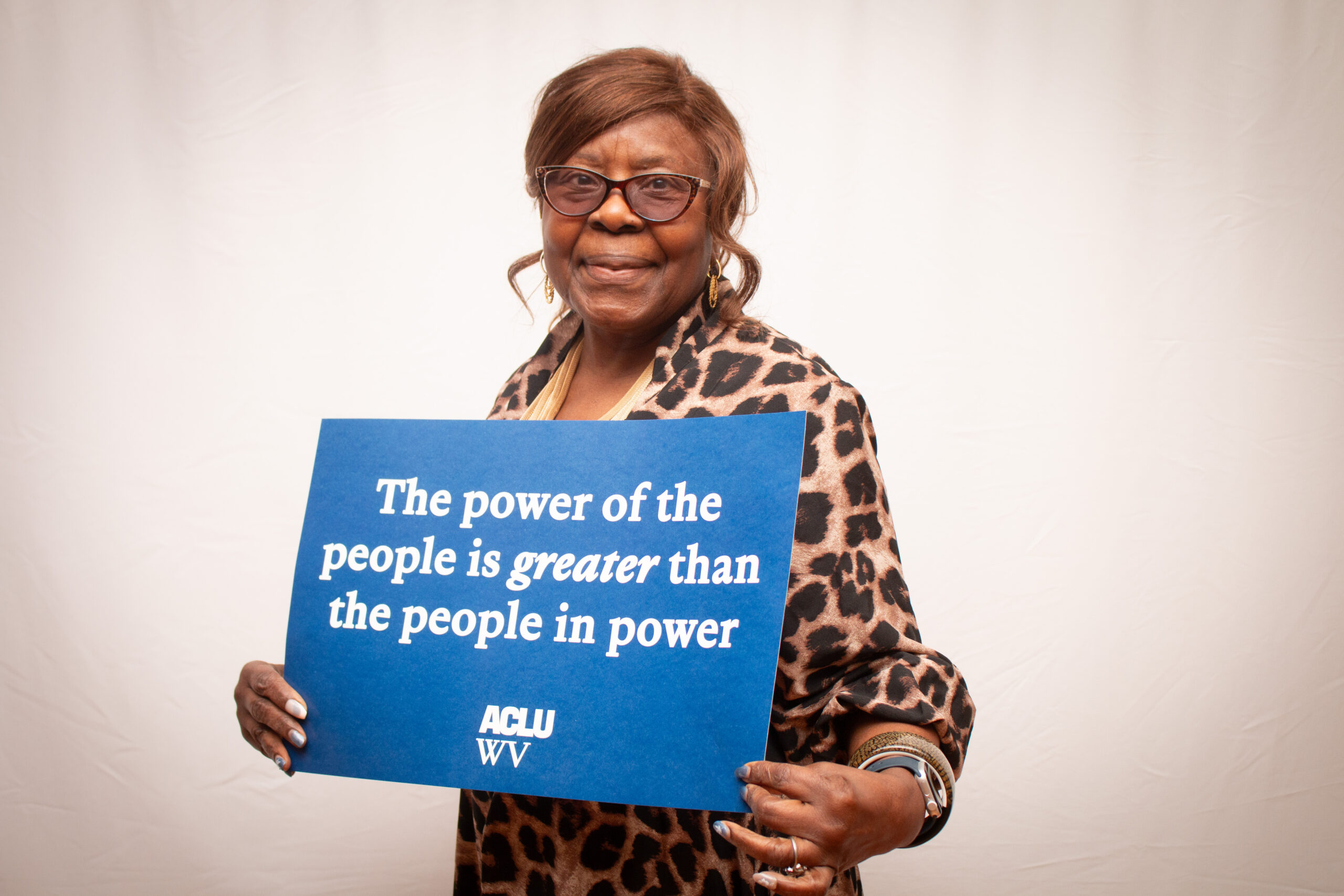 An older person wearing glasses, a leopard-print blouse, and gold jewelry holds a blue ACLU WV sign reading "The power of the people is greater than the people in power" against a light backdrop. They smile warmly at the camera.