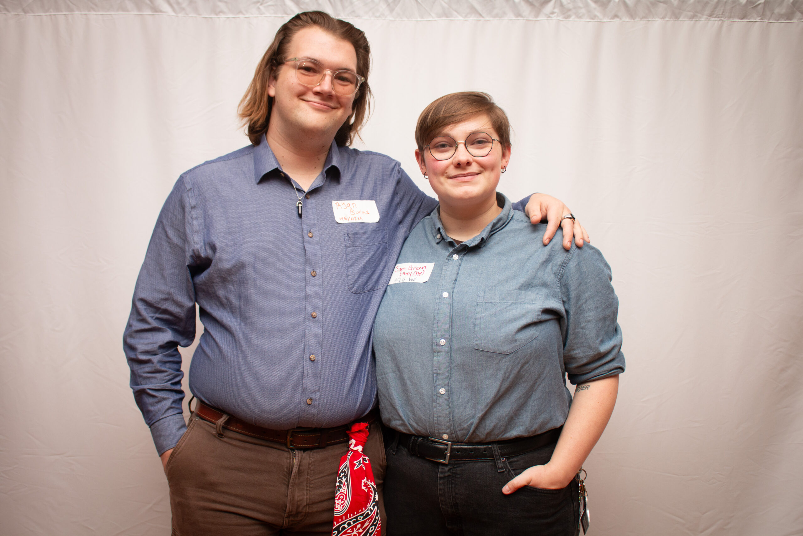 Two people wearing matching blue chambray shirts stand with arms around each other against a light backdrop, both wearing glasses and name tags. The person on left has shoulder-length hair and wears a belt, while the person on right has short hair with an undercut and wears a black belt. A red bandana is visible on the left figure.
