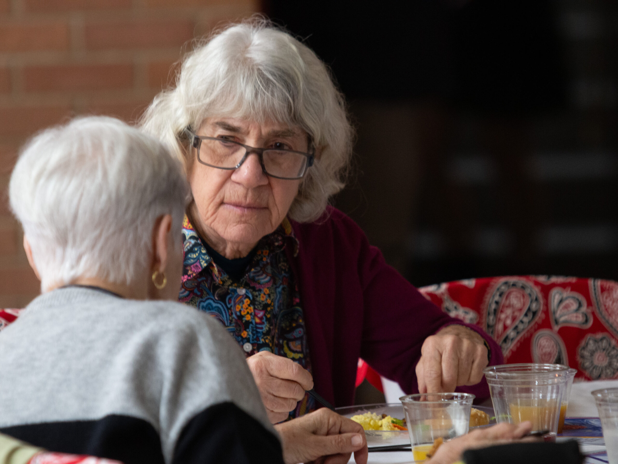 An older person with white hair and glasses sits at a dining table engaged in conversation with someone whose back is to the camera. They wear a maroon cardigan over a colorful paisley scarf and have food and drinks on the table. Red patterned decorations are visible in the background.