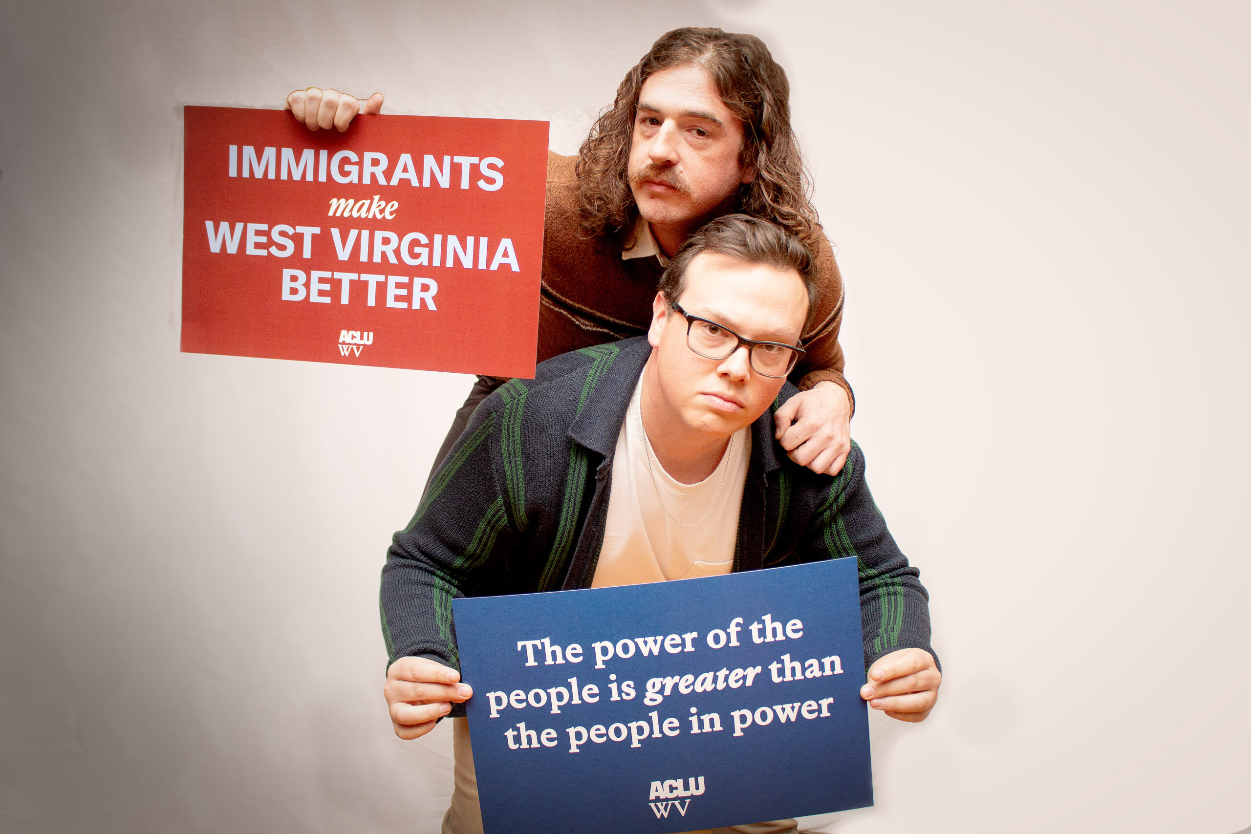Two people pose seriously holding advocacy signs against a white backdrop. The person standing behind holds a red sign reading "IMMIGRANTS make WEST VIRGINIA BETTER" (ACLU WV), while the person in front holds a blue sign reading "The power of the people is greater than the people in power" (ACLU WV). Both wear dark clothing.